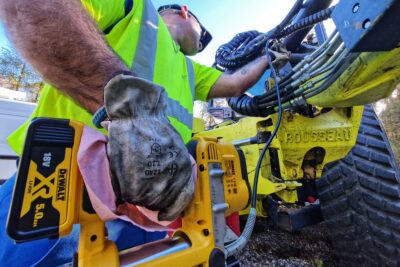 La photo est prise en contre plongée. Sur la partie gauche un homme en vêtement de travail jaune fluo et bleu électrique, casquette vissée sur la tête, tient dans sa main droite un appareil jaune relier par un petit tuyau noir à une espèce de tige qu'il tien dans sa main gauche. En arrière plan un imposant tracteur jeune vert dont on ne distingue que des cables hydrauliques entournés de gaines noires. L'hommes est en train de graisser à l'aide de la tige les mécanismes hydrauliques. - Agrandir l'image 5 sur 7, fenêtre modale