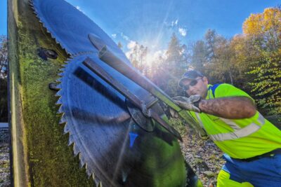 La photo est prise en contre plongée. Sur sa partie gauche, gros plans sur des disques de métal aux dents acérées sont fixés sur ce qui ressemble à une plaque de métal jaune. Un homme en vêtement de travail jaune fluo et bleu électrique est en train de les fixer sur la plaque à l'ai d'un outil en métal composé de leviers métallique. Un rayon de soleil s'invite au milieu de la photo. Le ciel est bleu parsemé de quelques nuages. - Agrandir l'image 4 sur 7, fenêtre modale
