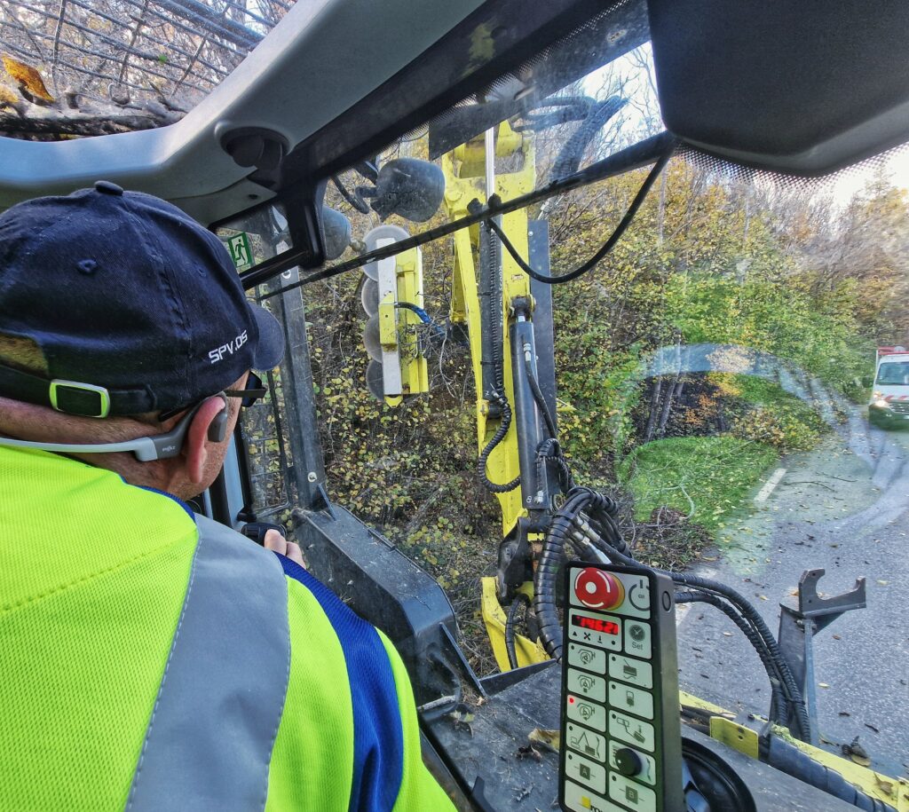 La photo a été prise à l'intérieur de l'habitacle d'un tracteur. Au premier plan, un homme nous fait dos. Il porte un vêtement de travail jaune fluo et bleu électrique et une casquette. On devine qu'il manipule un joystick qui actionne le bras mécanique articulé que l'on voit à l'extérieur, au travers de la vitre du tracteur. Une rangée de disques aux dents tranchantes se trouve au bout de ce bras mécanique. Il est en train de découper les branches dépassant sur la chaussée des arbres et buissons qui se trouve au bord de la route. - Agrandir l'image, fenêtre modale