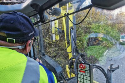 La photo a été prise à l'intérieur de l'habitacle d'un tracteur. Au premier plan, un homme nous fait dos. Il porte un vêtement de travail jaune fluo et bleu électrique et une casquette. On devine qu'il manipule un joystick qui actionne le bras mécanique articulé que l'on voit à l'extérieur, au travers de la vitre du tracteur. Une rangée de disques aux dents tranchantes se trouve au bout de ce bras mécanique. Il est en train de découper les branches dépassant sur la chaussée des arbres et buissons qui se trouve au bord de la route. - Agrandir l'image 3 sur 7, fenêtre modale