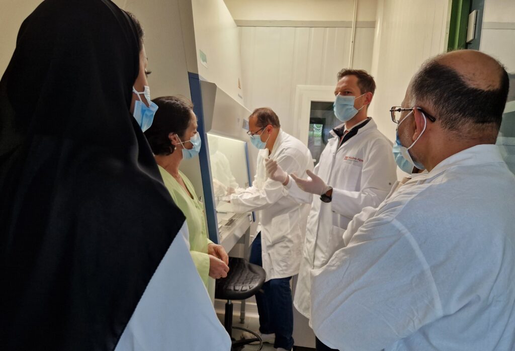 La scène se passe dans un laboratoire. On y voit 5 protagonistes, tous sont habillés de blouses de laboratoire et tous portent des masques de protection couvrant leur nez et bouche. Au fond, sous une hotte d'analyse, une homme est en train de manipuler quelque chose sous la hottes. Au premier plan, les quatre autres personnage, deux femmes et deux hommes sont en train d'échanger. - Agrandir l'image, fenêtre modale