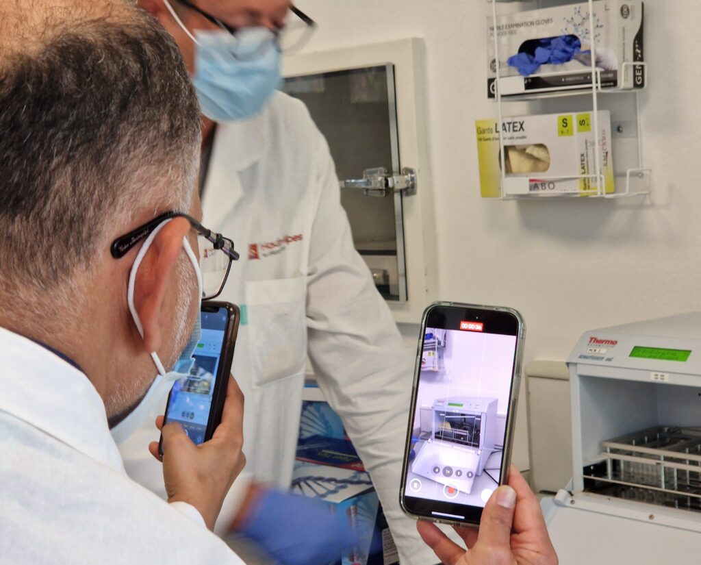 Au premier plan, un homme de dos dont on ne voit que la tête. Il porte son regard sur un écran de téléphone en train de filme un machine carré de laboratoire qui ressemble à une espèce de petit four. C'est l'homme du premier plan qui est en train de filmer la machine. À côté de lui, un autre téléphone est en train de filme. En arrière plan, en flou, un autre homme. La scène se passe dans un laboratoire. Les protagonistes portent des blouses blanches. - Agrandir l'image, fenêtre modale