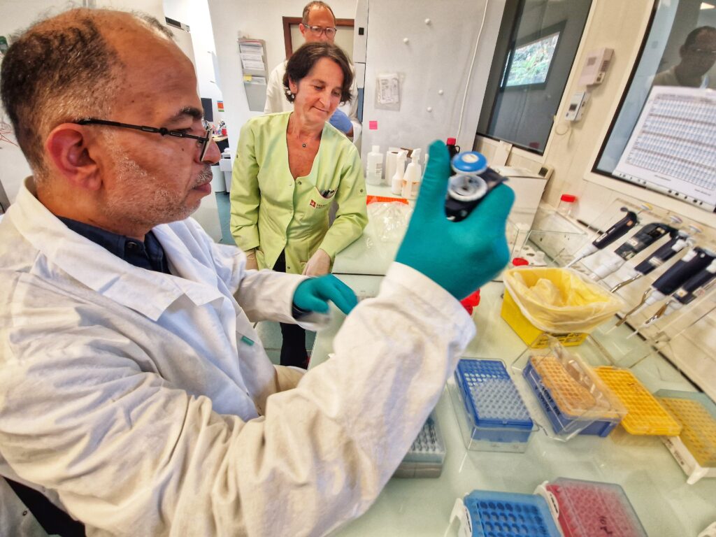 La scène se passe dans un laboratoire. Au premier plan, sur la partie gauche de la photo, un homme en blouse blanche, crâne dégarni, lunettes sur le nez. Il porte des gants vert en latex. Dans sa main droite une pipette électrique. Devant lui, posés sur un plan de travail en verre, une multitude de petits boitiers rectangulaire munis de petits trous. À côté de l'homme, une femme, cheveux courts et habillée d'une blouse vert pâle pose son regard sur le plan de travail. Encore derrière elle, on devine un autre homme, en blouse blanche qui lui aussi regarde le plan de travail. - Agrandir l'image, fenêtre modale