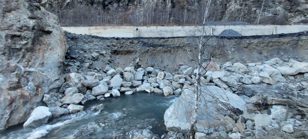 Photo prise depuis le lit d'une rivière donnant sur une berge en contre bas d'une route. La berge s’effondre emportant avec elle la route. Des blocs rocheux ont été positionné pour tenté de stopper le phénomène. En surplomb, un ouvrage d'art en béton gris, une galerie. - Agrandir l'image, fenêtre modale