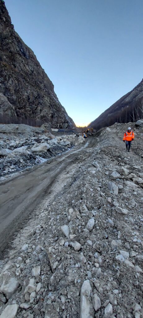 Au fond d'une vallée encaissée des gravats de terre et blocs rocheux, couleur grise. Une homme en habit de chantier orange fluo marche sur la droite. Au dernier plan, on devine des engin de chantier. - Agrandir l'image, fenêtre modale