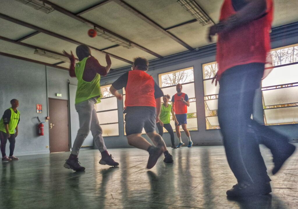 Un groupe de personnes, chasubles jaunes pour les une, orange pour les autres, en train de jouer au hand ball dans un gymnase. - Agrandir l'image, fenêtre modale