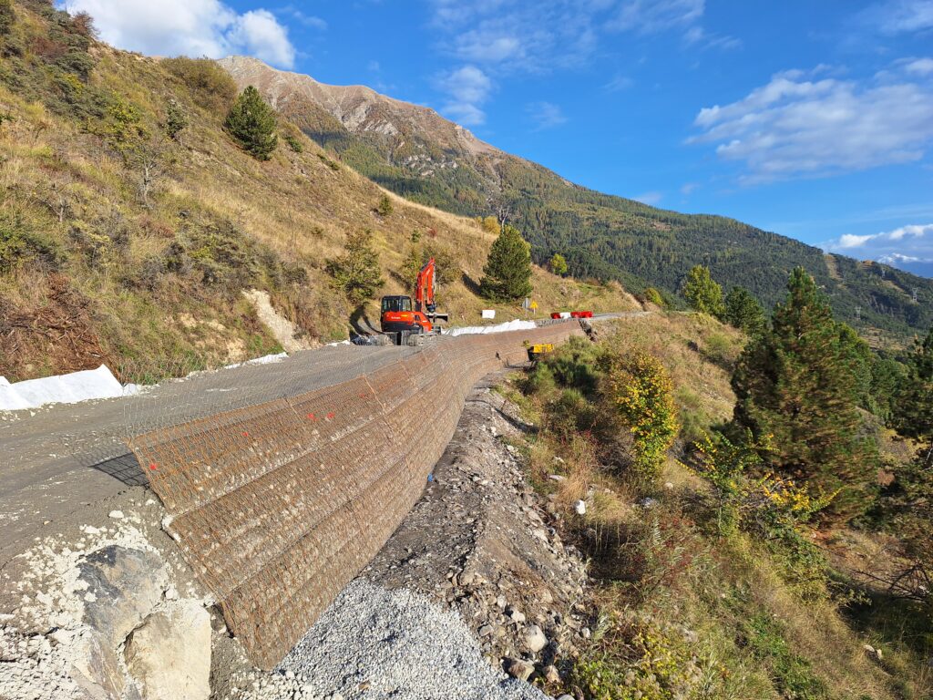 La photo est divisé en deux. Un route en travaux sépare les deux partie dans le sens de la longueur. Sur la route, se situe sur une pente soutenue par des grillages sur sa partie droite. En son milieux une pelleteuse rouge est en train de travailler. Au dessus de la route, une pente montagneuse recouverte de végétation aux couleurs de l'automne. Idem en contre-bas de la route. Au loin des paysages vallonnés de montagne. Le ciel est bleu parsemé de nuages. - Agrandir l'image, fenêtre modale