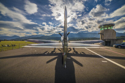 En plein milieu de la photo, sur une piste de décollage en bitume noir, un avion de la second guerre mondial de dos. La lumière du soleil reflète sur la carlingue. En second plan sur la droite, une tour de contrôle. Au bas de cette dernière est stationné une voiture de gendarmerie, type SUV. À gauche de l'avion, une partie herbacée fraichement tondue. Au loin les montagnes. Le ciel est bleu parsemé de nuages blancs . - Agrandir l'image, fenêtre modale