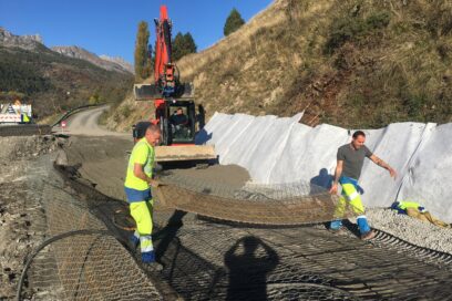 La scène se passe sur le chantier d'une route en reconstruction. Au milieu de la photo deux homme en vêtement de travail jaune fluo et bleu électrique portent une espèce de grillage métallique le plusieurs mètres de large. Grillage qui recouvre le sol. En dessous on aperçoit du remblai couleur gris. Derrière les deux hommes, une pelleteuse rouge pourvue, au bout de son bras mécanique articulé d'un godet noir en train d'aplanir du remblai déposé également par dessus ce grillage. En arrière plan, le reste de la route déjà existante et les montagne. - Agrandir l'image, fenêtre modale