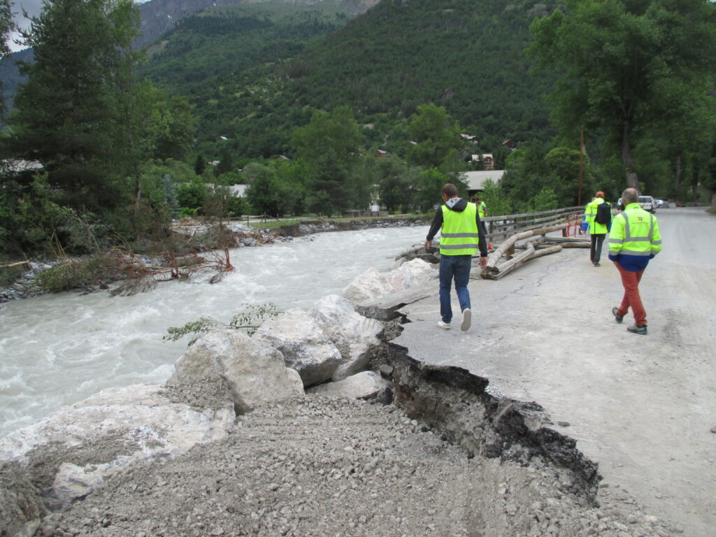 Des homme en blouson jaune fluo et bleu marche le long d'une rivière en furie. La berge est une route dévastée. - Agrandir l'image, fenêtre modale