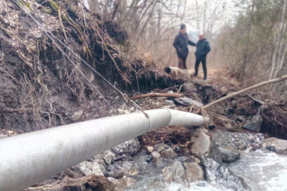 Au premier plan, la berge effondrée d'une rivière et une tuyau métallique qui ne tien que par un fil en acier. En arrière plan, dans le flou, deux homme habillé des vêtements chaud et sombre semblent constater les dégâts depuis la berge. Ils sont entouré de rivière. Le ciel est bas, la luminosité truste. - Agrandir l'image, fenêtre modale