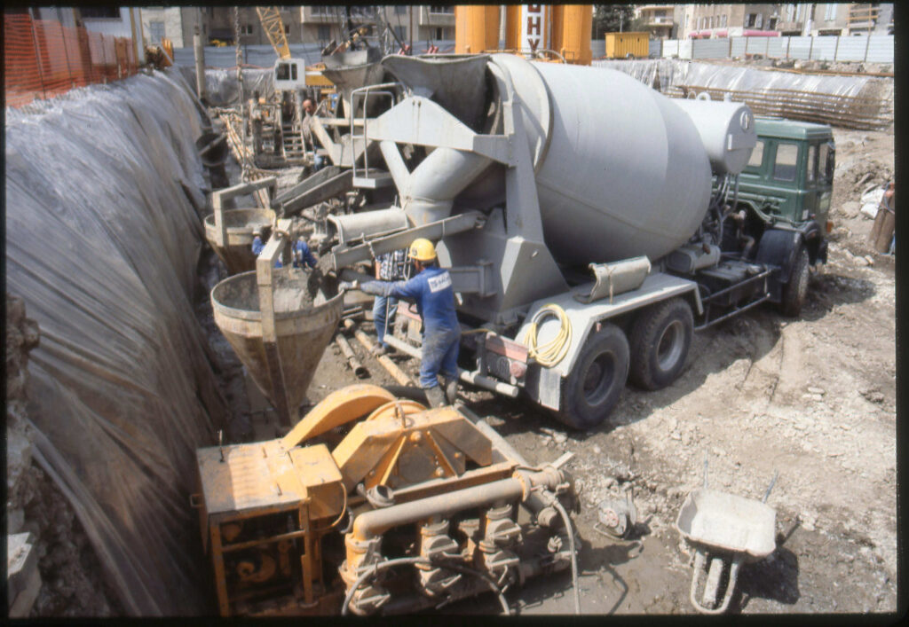 Au centre de la photos deux camions bétonnières au fond d'un trou béant où se tient le chantier déverse leur béton dans de grands cônes an métal. Des hommes en habit de chantier bleu surveillent que les opérations se déroulent bien. - Agrandir l'image, fenêtre modale