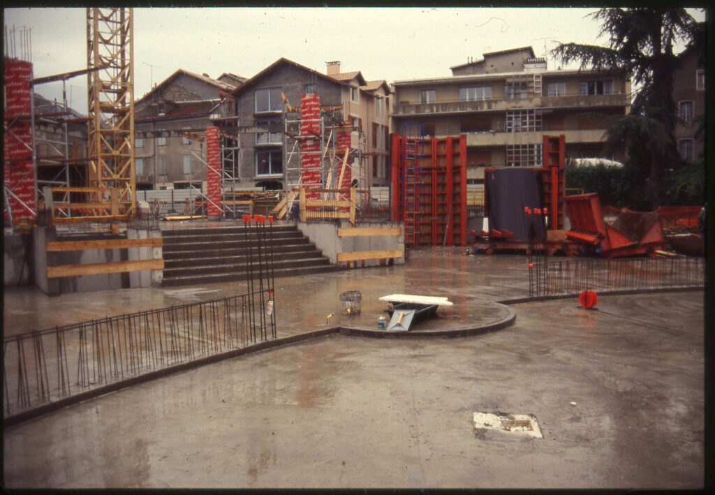 Un parterre en béton, quelques ferraillages qui sortent de terre, un escalier, lui aussi en béton, une crue, des colonnes encore emmaillotées, en arrière plan des maison de centre ville. La photo semble ancienne. - Agrandir l'image, fenêtre modale