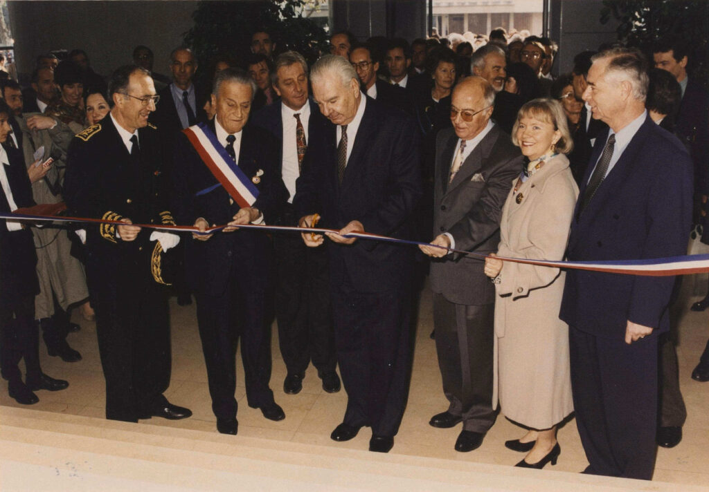 La photo semble ancienne. Une rangé d'homme en costume dont l'un porte une écharpe tricolore et à son côté un autre porte le costume d’apparat de Préfet. Celui au milieu, les cheveux gris, la mine toute en rondeur est une train de couper un ruban inaugural. Derrière eux une foule nombreuse. La scène sa passe dans la hall de l’Hôtel du Département. La photo date du 28 novembre 1994. - Agrandir l'image, fenêtre modale