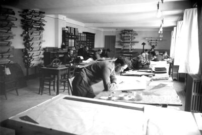 Une photo en noir et blanc qui date du début du XIXè siècle. Debout mais accoudés sur leurs bureau des hommes planche sur des plans. Sur leurs bureau on peut voir des équerres, des livres. Sur les murs retenu par des crocher en métal noir incurvés des plans enroulés sur eux mêmes. - Agrandir l'image 7 sur 7, fenêtre modale