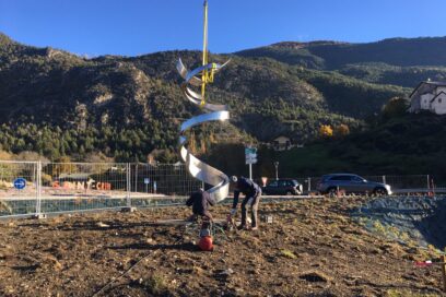 Deux hommes sont en train d'installer la sculpture monumentale en extérieur. Au deuxième plan, nous apercevons le rond-point près de l'aérodrome de Saint-Crépin. En arrière plan, les montagnes. - Agrandir l'image, fenêtre modale