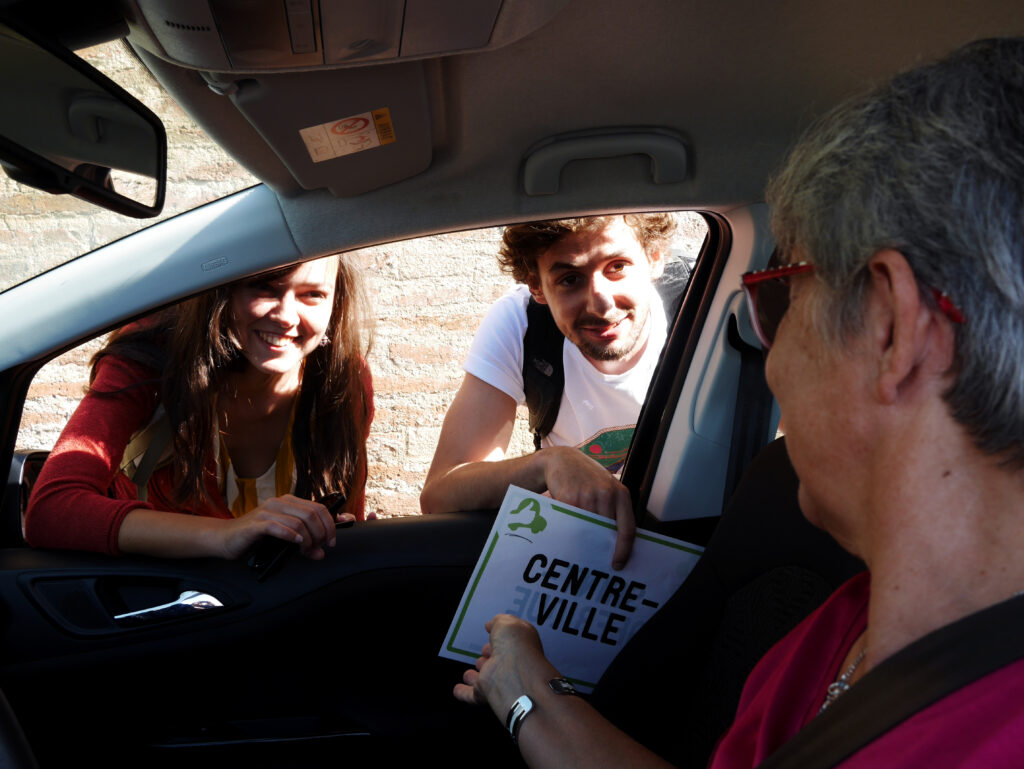 La photo est prise de puis l'habitacle d'une voiture. Au premier plan, une dame les cheveux grisonnants et vêtue d'un t-shirt rose est assise place conducteur. Elle tourne la tête vers deux jeunes, une fille au cheveux long et d'un cardigan ocre et un garçon cheveux bouclés et t-shirt blanc, accoudés à l'extérieur du véhicule. Les trois personnages sourient. Le garçon a entre les mains une pancarte sur laquelle on peut lire centre-ville. On devine que la dame va prendre en stop les deux jeunes. - Agrandir l'image, fenêtre modale