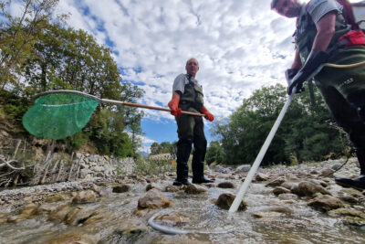 Deux homme habillés de waders et des gants en plastique épais se tiennent debout au milieux d'une rivière. L'un est équipé un anneau électrique situé au bout d'une perche. L'autre d'une grande épuisette au maillage vert. - Agrandir l'image 3 sur 6, fenêtre modale