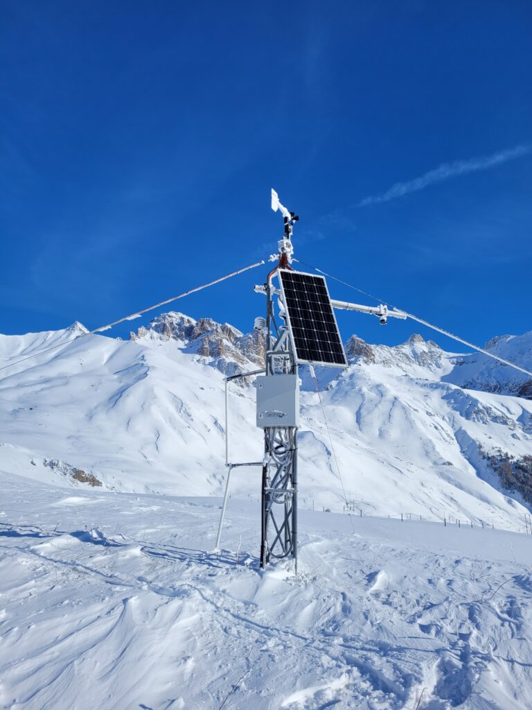 Le paysage est blanc de neige, en grande quantité. Au fond on distingue des montagnes enneigées. Le ciel est bleu sans nuages. Au milieu, un mât métallique mini d'une armoire électrique, d'un panneau solaire et d'une girouette au sommet. - Agrandir l'image, fenêtre modale