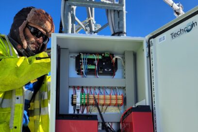 Au milieu de la photo une armoire électrique accrochée à un mât métallique. À l'intérieur une multitude de composant électronique et deux batterie rouge. À l'arrière une homme en vêtement et veste de travail jaune fluo et bleu électrique, chapka marron sur la tête, lunette de soleil vissée sur le nez. Il porte également un tour de cou marron et des guêtres en nylon. Il est visiblement en train de réparer quelque chose à l'arrière de l'armoire électrique. Il semble y avoir un importante couche de neige au sol. Le ciel est bleu. Sou l'armoire électrique, une multitude de câbles protégé de geines grise en plastique. - Agrandir l'image, fenêtre modale