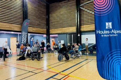 Dans un gymnase au parquet clair, des personne en fauteuil roulant réuni en cercle en train de partager leur expérience. Au premier plan de la photo, un oriflamme bleu marine sur lequel on peut lire Hautes-Alpes. - Agrandir l'image, fenêtre modale