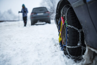 Au premier plan, gros plan sur une roue de voiture équipée de chaine à neige. En second plan, en flou, une voiture immobilisée et une personne emmitouflée qui marche avec des bâtons. Et de la neige partout. - Agrandir l'image, fenêtre modale