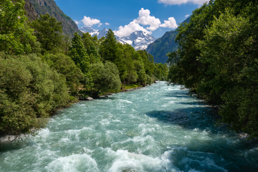 Au milieu de la photo une rivière aux eaux vives couleur bleu vert. Sur les berges, des forêts aux arbres verdoyant. Au loin, des montagne avec quelques névés. Le temps est au soleil. - Agrandir l'image, fenêtre modale