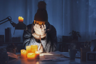 Une femme en bonnet, écharpe et gant se tient le visage entre les mains. Il semble faire froid dans son logement éclairé à la seule lueur de quelques bougies. - Agrandir l'image, fenêtre modale