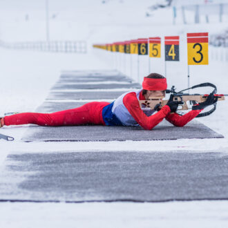 Dans un paysage blanc de neige, un biathlète habillée d'une tenue près du corps rouge est allongée sur un tapis gris. Elle est en position de tir couché. On distingue d'autre tapis gris au sol qui se succèdent en une multitude de couloirs délimités par un piquet (un par couloir) surmonté d'un rectangle en métal portant un chiffre