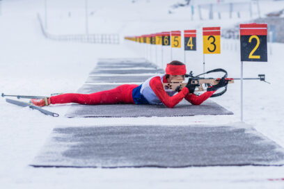 Dans un paysage blanc de neige, un biathlète habillée d'une tenue près du corps rouge est allongée sur un tapis gris. Elle est en position de tir couché. On distingue d'autre tapis gris au sol qui se succèdent en une multitude de couloirs délimités par un piquet (un par couloir) surmonté d'un rectangle en métal portant un chiffre - Agrandir l'image, fenêtre modale