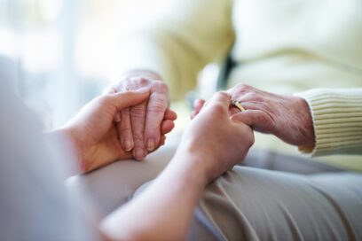 Gros plan sur deux paire de mains qui se tiennent. L'image laisse suggérer que deux personnes sont assises l'un en face de l'autre et se tiennent les mains. On imagine aussi qu'il s'agit d'une personne plus jeune et l'autre plus âgée. La photo est prise depuis le point de vue de la personne la plus jeune. En face la dame au mains d'un certain âge porte un pull jaune pâle et un pantalon gris. - Agrandir l'image, fenêtre modale
