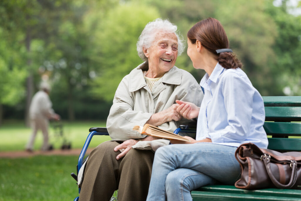 Deux femmes assises sur un banc dans un parc. Elles se tiennent la main. L'un est jeune, brune, habillée d'une chemise bleu pâle. L'autre est plus âgée, les cheveux courts et blancs. Elle porte une vêtes légère couleur beige, un pantalon marron et un pull jaune pale. Un large sourire illumine son visage - Agrandir l'image, fenêtre modale