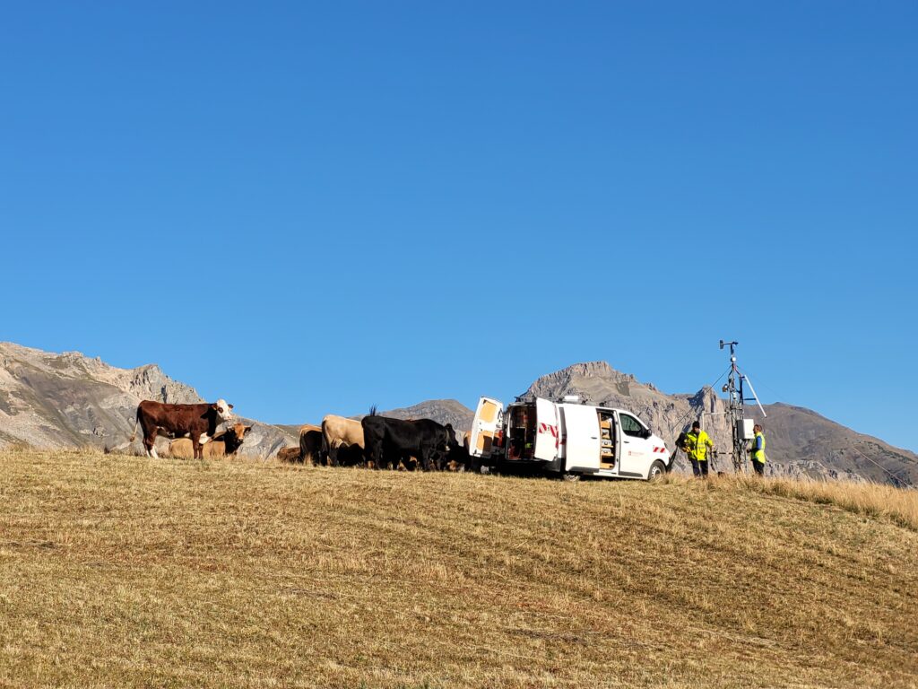 Au milieu de la photo, un troupeau de vaches noirs, marron foncé et couleur sable. Elles sont juste à côté d'une camionnette blanche dont les porte sont ouvertes. À l'intérieur on distingue comme des étagères remplis d'outils divers d'électricien. Dans le prolongement, deux hommes se tiennent débout, habillés en vêtement de travail jaune fluo et bleu électrique. Dérrière eux se dresse un mât métallique compose d'une armoire électrique à mi-hauteur et d'une girouette au sommet. Sur la partie inférieur de la photo, de l'herbe sèche jaunie. Au fond, on aperçoit des montagnes sans végétation. La partie supérieure de la photo, qui occupe presque la moitié de la photo est composée du ciel qui est bleu et sans l'ombre d'un nuage. - Agrandir l'image, fenêtre modale