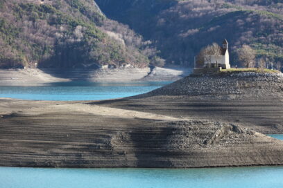 Au milieu d'un lac (Serre-Ponçon), un îlot formé de deux monticules. Sur le plus imposant des deux, à droite, une petite chapelle (Saint-Michel) entourée de verdure. Tout autour de l'îlot, de l'eau couleur bleu-vert. En arrière plan, des montagnes au flans recouverts de forêt. Les feuillu sont dénudés, les conifères apportent ici et là des touches de vert dans un paysage plutôt gris. Le temps est au soleil. - Agrandir l'image, fenêtre modale