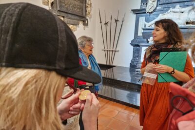 Au premier plan une dame blonde, casquette noir sur la tête et portant des lunettes à la monture rouge regarde de très près une médaille dorée représentant le profil de François de Bonne de Lesdiguières. Au second plan, une jeune quide du musée en rose orange, portant une pochette cartonnée verte des ses bras et un sachet en plastique contenant un autre fac-similé de la médaille de François de Bonne de Lesdiguières et en train de discuter avec une femme d'un certain âge, cheveux court et gris portant des lunettes rosés, une doudoune rose un pull beige. Les deux femmes discutent en souriant. en arrière plan on aperçoit le mausolée principalement en marbre noir de mausolée Lesdiguières. - Agrandir l'image 9 sur 13, fenêtre modale
