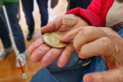 En gros plan une main tenant en son sein une médaille dorée représentant François de Bonne de Lesdiguières. L'autre main de la personne tenant cette médaille la parcourt du bout du doigt. En arrière plane on aperçoit les jambes et cannes de locomotion des personnes qi se tiennent derrière. - Agrandir l'image 8 sur 13, fenêtre modale