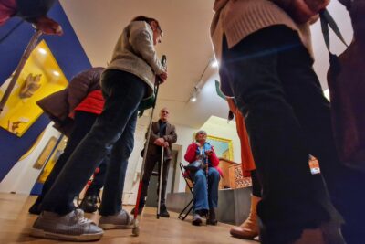 Une groupe de personnes aveugles et malvoyantes, cannes de locomotion en main sont réunies autour de la guide du musée leur donnant des explications. Au centre de la photo, une dame en doudoune rose et jeans est assise sur une chaise pliante. - Agrandir l'image 7 sur 13, fenêtre modale