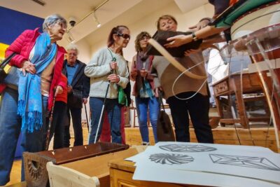 En premier plan des coffres et berceaux en bois typiques du Queyras. Dessus, sont posées des feuilles reproduisant des rosaces du Queyras via thermo gonflage. Au deuxième plan, un groupe de visiteurs malvoyants écoute les explications d'une jeune guide du musée qui leur présente un cartable en bois foncé du Queyras. En arrière plan on devine du mobilier ancien du Queyras. - Agrandir l'image 6 sur 13, fenêtre modale