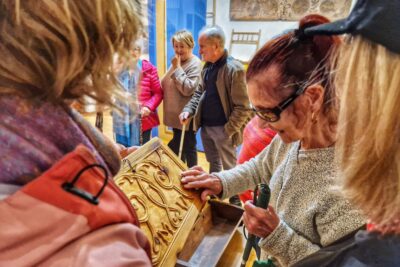 Une femme aveugle, lunette de soleil sur le nez et canne de locomotion dans une main touche de sa main libre les détails en rosace du fac-similé d'un coffre du Queyras. En arrière plan des visiteurs s'attardant sur une autre oeuvre du musée. - Agrandir l'image 4 sur 13, fenêtre modale