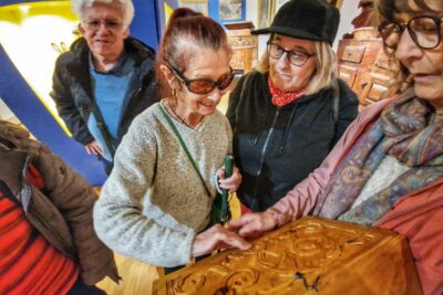 Une dame aveugle, lunette de soleil sur le nez et canne de locomotion dans une main explore de sa main libre le fac-similé d'un coffre du Queyras. À coté d'elle, un groupe de personnes malvoyantes attendent leur tour. La scène se passe dans l'une des salle du Musée muséum départemental des Hautes-Alpes. - Agrandir l'image 3 sur 13, fenêtre modale