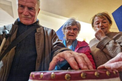 Un homme malvoyant explore du bout des doigts un tambour de coutrière du Queyras. À coté de lui, deux femmes les yeux rivés sur l'objet. La scène se passe dans l'une des salle du Musée muséum départemental des Hautes-Alpes. - Agrandir l'image 2 sur 13, fenêtre modale