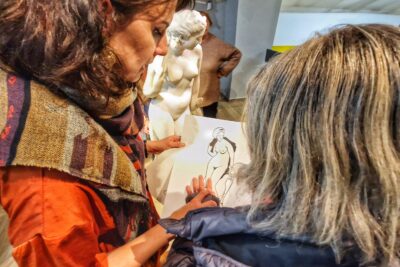 Au premier plan, une jeune femme voyante guide la main d'une personne aveugle sur une feuille représentant la statue de Léda et Zeus en forme de signe. Statue qui se retrouve au deuxième plan au centre de la photo. - Agrandir l'image 13 sur 13, fenêtre modale